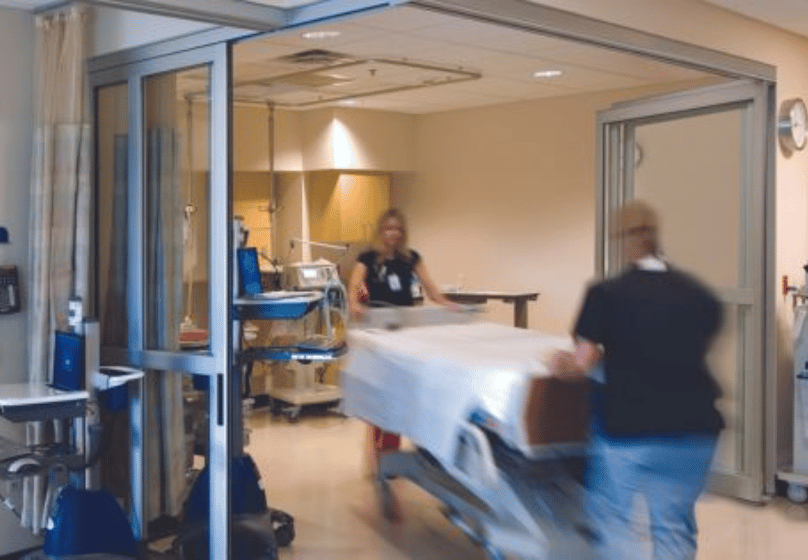 Two medical staff members are moving an empty hospital bed through a hallway in a healthcare facility, passing by commercial doors maintained with reliable automatic door service.