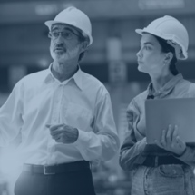 Two people wearing hard hats stand indoors; one holds a laptop while the other gestures, suggesting a discussion or inspection about commercial door installation at a work site.