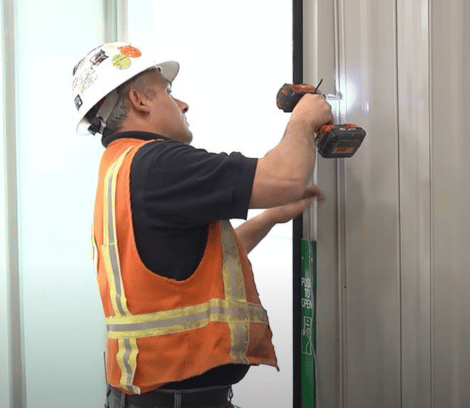 A construction worker in an orange safety vest and hard hat uses a power drill to install or secure a metal panel indoors, preparing the space for commercial doors as part of a larger commercial door repair project.