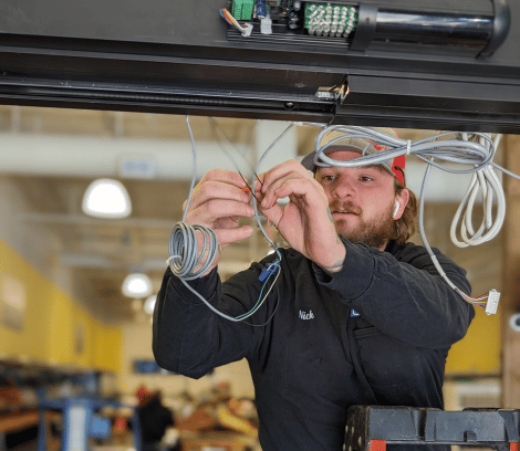 A man in work attire connects wires to an overhead device inside an industrial building, performing automatic door maintenance. Shelves and lights are visible in the background.