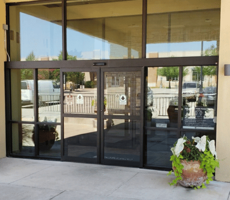 Glass automatic sliding doors of a building entrance, expertly crafted through professional commercial door installation, with a potted plant to the right and visible reflections of parked cars and trees.