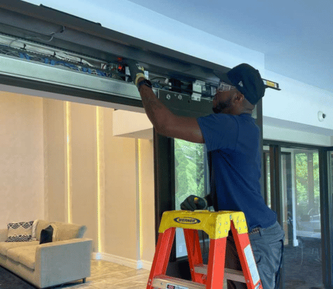 A person in a blue shirt and cap stands on a ladder, working with wiring and equipment inside the top frame of a sliding glass door in a modern living space, performing automatic door repair to ensure seamless operation.