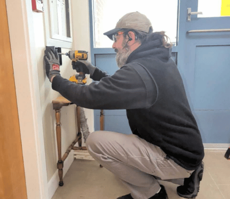 A person wearing gloves and a cap uses a power drill to install or repair an electrical outlet on an indoor wall near a blue door, possibly as part of commercial door installation or automatic door maintenance.