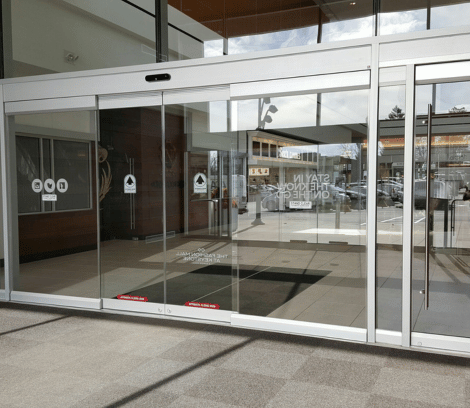 Automatic glass sliding doors at the entrance of a modern building, leading to a lobby area with reflections on the glass and a view of the parking lot outside. These commercial doors provide both convenience and style, ensuring smooth entry while professional automatic door service keeps them operating flawlessly.