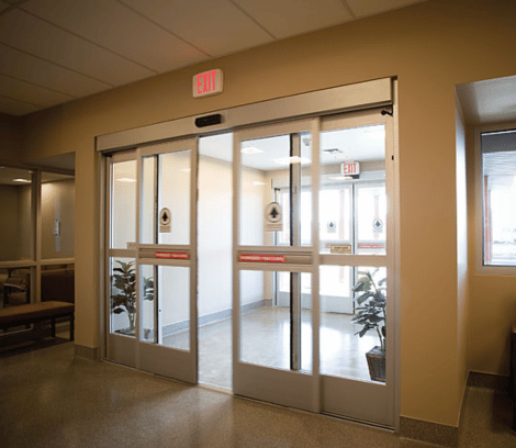 Automatic sliding glass doors in a building hallway, with EXIT signs above and potted plants visible on both sides of the entrance—an inviting space maintained through professional automatic door service to ensure seamless and reliable operation.
