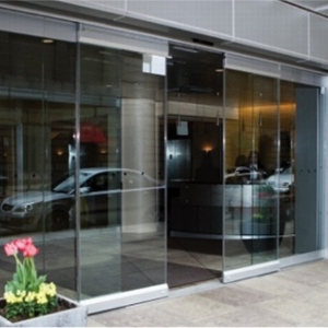 Glass sliding doors at a building entrance lead to a lobby interior. Outside, a car is parked and a small planter with pink flowers brightens the scene. Nearby, an automatic door service vehicle stands ready for any commercial door installation or repair needs.